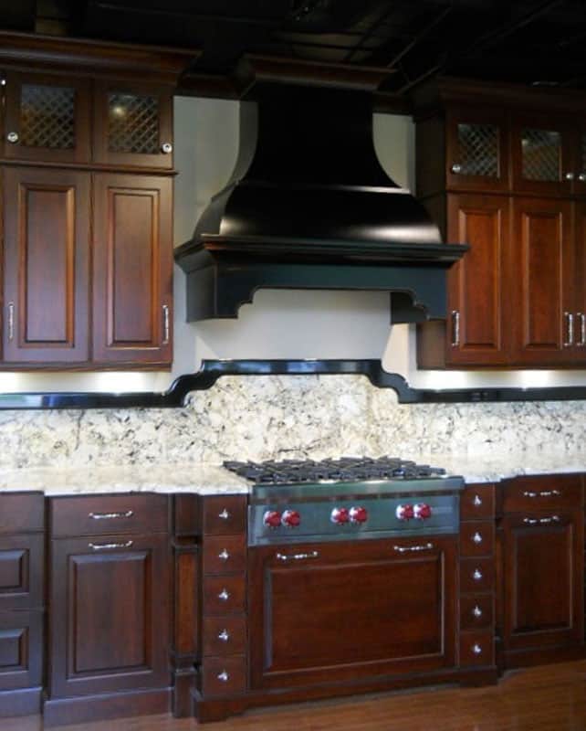 An architectural luxury kitchen design highlighted by a custom dark-stained D-Series wood range hood featuring sweeping lines, classic corbels, and a custom-scaled chimney.